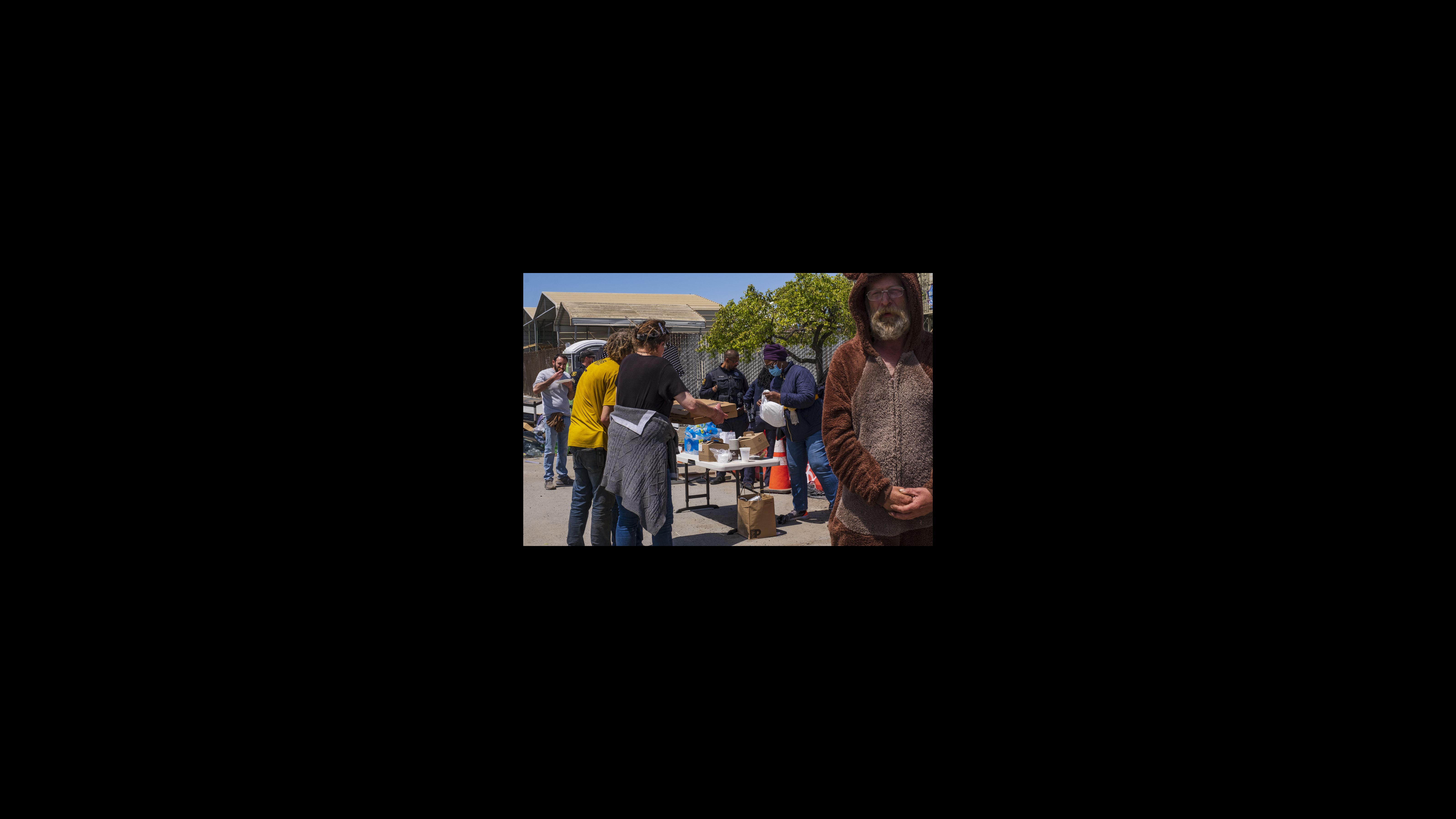 Radu orders a dozen pizzas for his crew, and they break for lunch. Second Street residents linger nearby and wait for leftover pizza. Moments later, Eva Ahmed offers slices of two pepperoni pizzas to more than a dozen residents. A woman with her back to the camera holds two pizza boxes as she and a few other people stand near a folding table that holds coffee and cups on it. A man wearing a brown one-piece bear-style outfit with a hood that has ears attached stands in the right foreground facing the camera.