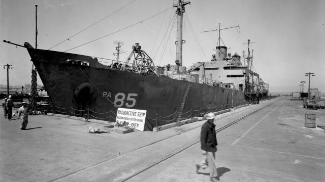 Hundreds of blue-collar shipyard workers were exposed to potentially dangerous amounts of radiation while attempting to “decontaminate” the USS Gasconade and other vessels hit by a 1946 atomic bomb test in the Pacific and brought to San Francisco for study. Source: National Archives and Records Administration. black-and-white historical photo of men walking on a dock in front of a naval ship, with a sign that says "radioactive ship, authorized personnel, keep off"