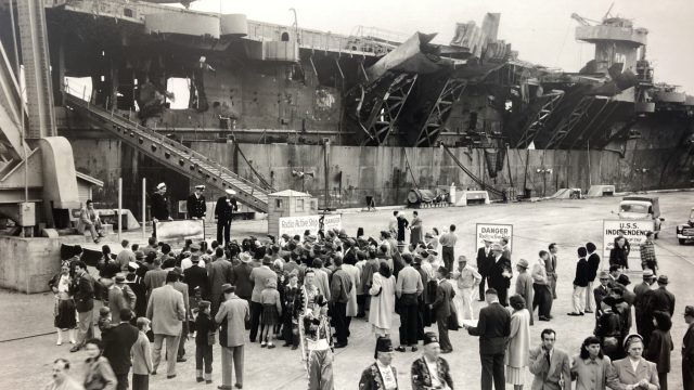 In 1947, the USS Independence — recently blasted by an atomic bomb test in the Pacific — went on public display at the Hunters Point Shipyard for its Navy Day celebration. It would end its service packed with other radioactive waste and scuttled just beyond the Golden Gate. Source: National Archives and Records Administration. a black-and-white photo of a large military ship with dozens of civilians gathered on a dock to admire, with a sign in the background saying "radio active ship - danger"